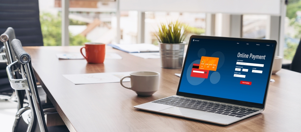 A laptop sitting on a desk with coffee, papers, and a plant. The laptop has the word online payment on it as someone makes a payment through a payment gateway.