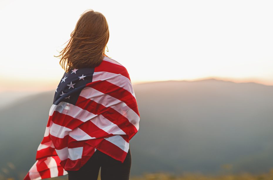 A woman wrapped in the United States flag is staring out at a mountain.